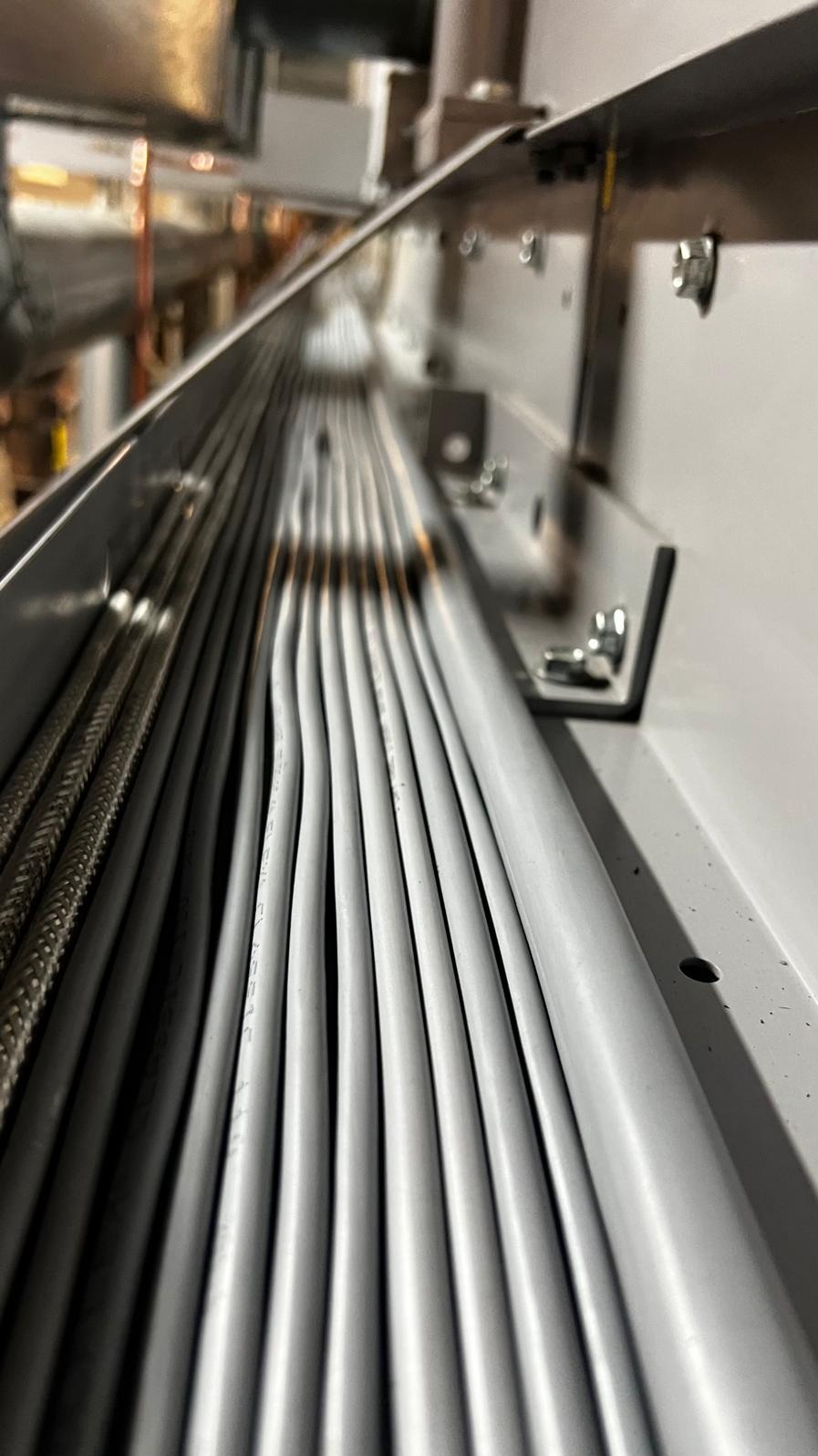 Równolegle ułożone szare przewody w metalowej korycie instalacyjnej, widoczne mocowania i elementy konstrukcyjne.