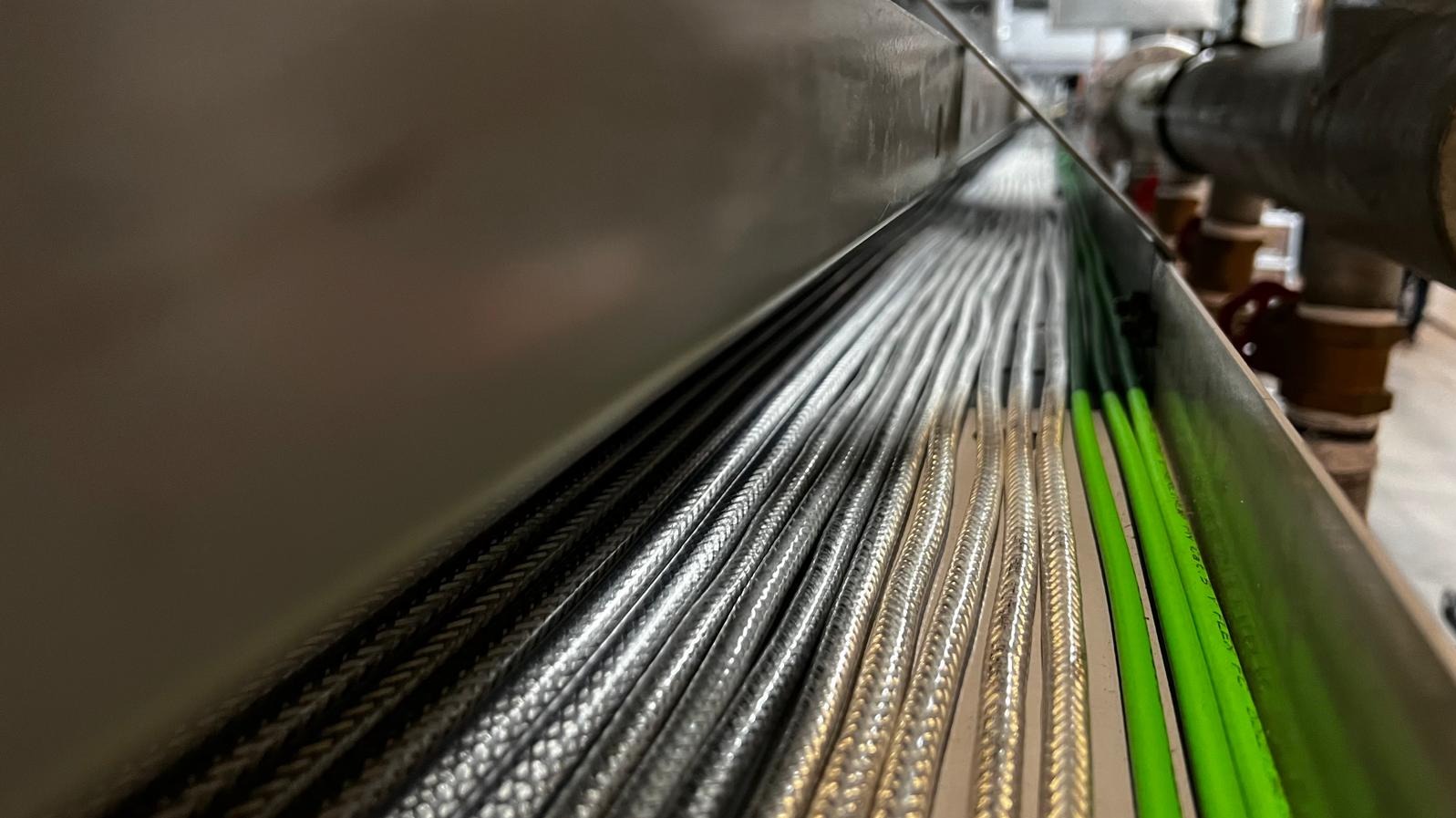 Metalowa korytka kablowa wypełniona równolegle ułożonymi przewodami w oplocie, część z nich w zielonej izolacji, widoczne rury instalacyjne w tle.