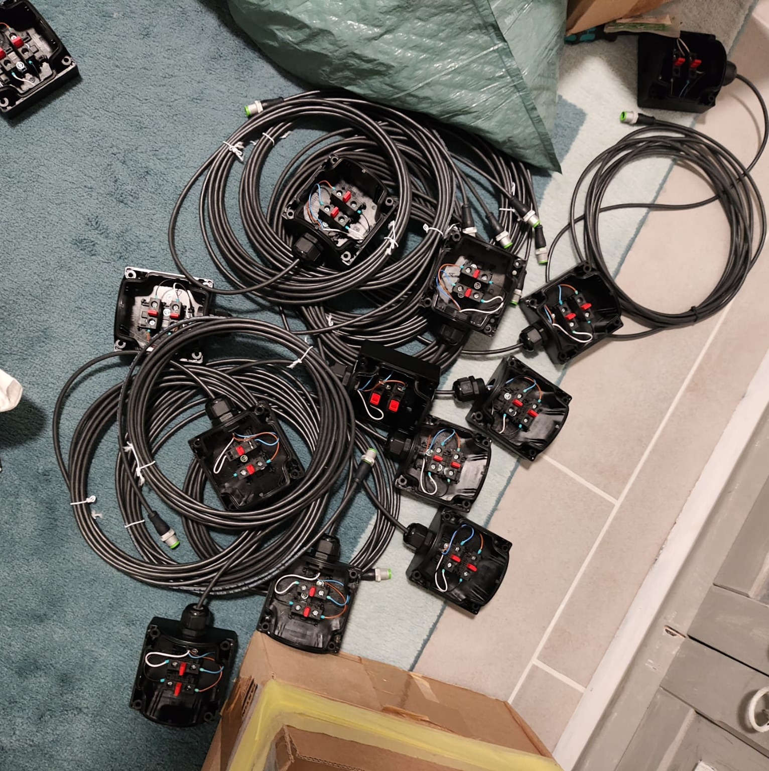 Przygotowane do montażu czarne puszki instalacyjne z podłączonymi przewodami na tle niebieskiego dywanu i szarej podłogi.