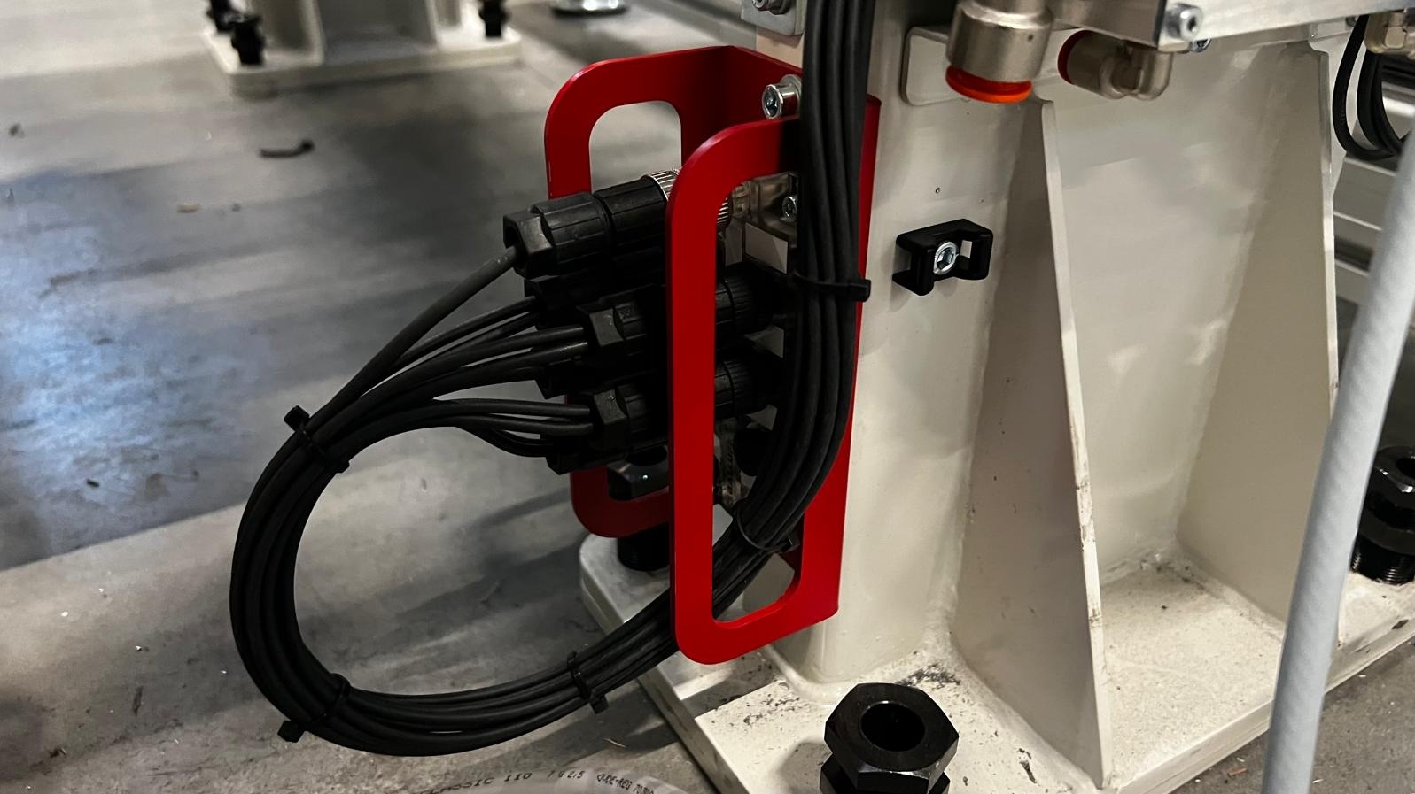 Zestaw podłączonych przewodów elektrycznych z czarnymi złączkami, zabezpieczonych czerwoną metalową ramką na białej konstrukcji urządzenia.