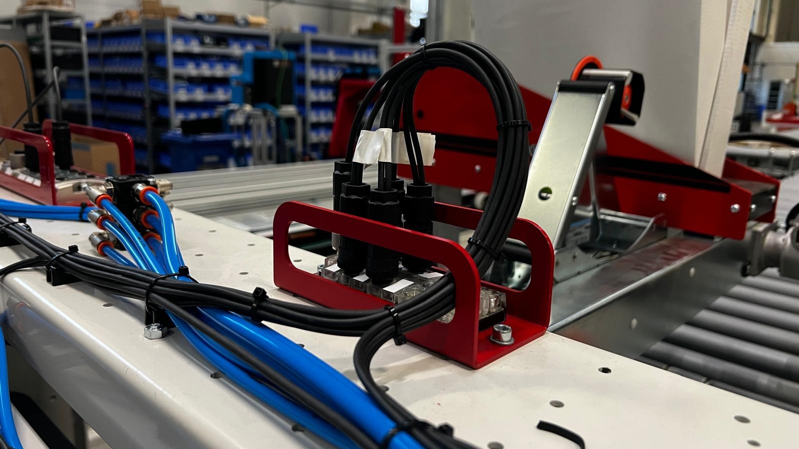 Detal instalacji elektrycznej: czarne kable przymocowane do przezroczystej listwy zaciskowej w czerwonej obudowie, niebieskie przewody pneumatyczne z pomarańczowymi złączkami, uporządkowane...