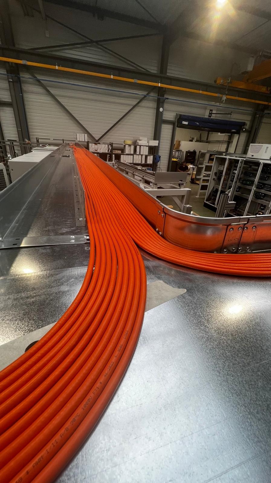 Równolegle ułożone pomarańczowe kable w metalowej korycie kablowej, widoczne napisy na kablach, tło warsztatowe z regałami i elementami maszyn.