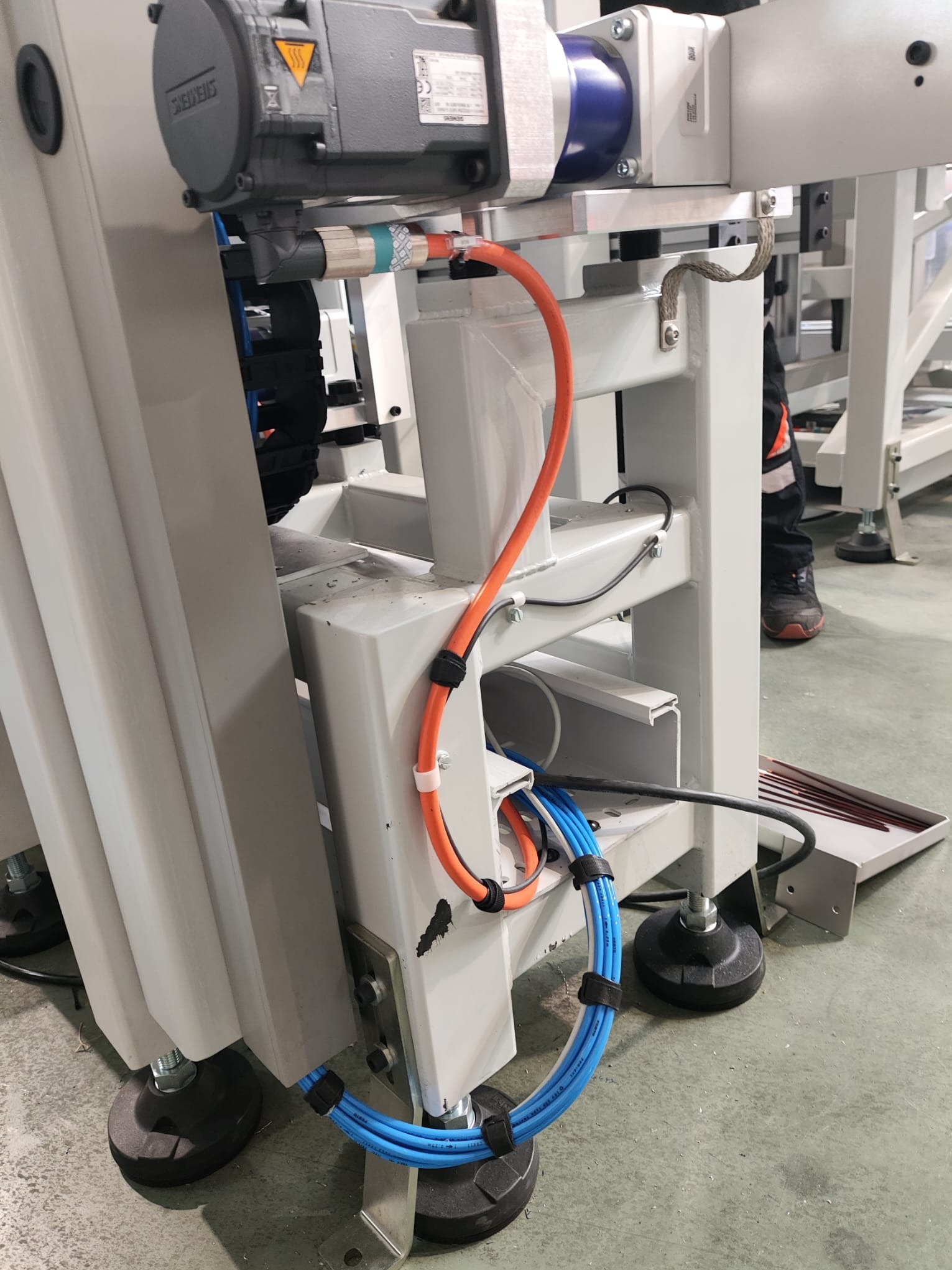 Szczegółowe ujęcie fragmentu maszyny przemysłowej z widocznymi przewodami elektrycznymi w kolorach pomarańczowym i niebieskim, przymocowanymi do metalowej konstrukcji za pomocą czarnych opasek...