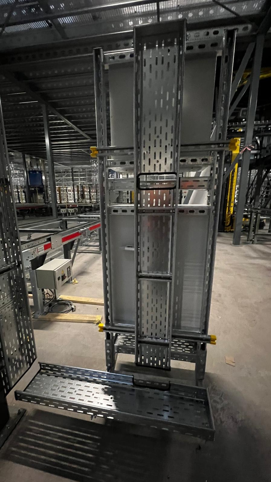 Metalowa konstrukcja nośna z perforowanymi elementami, prawdopodobnie do montażu tras kablowych w hali przemysłowej, widoczne elementy mocujące w kolorze żółtym.