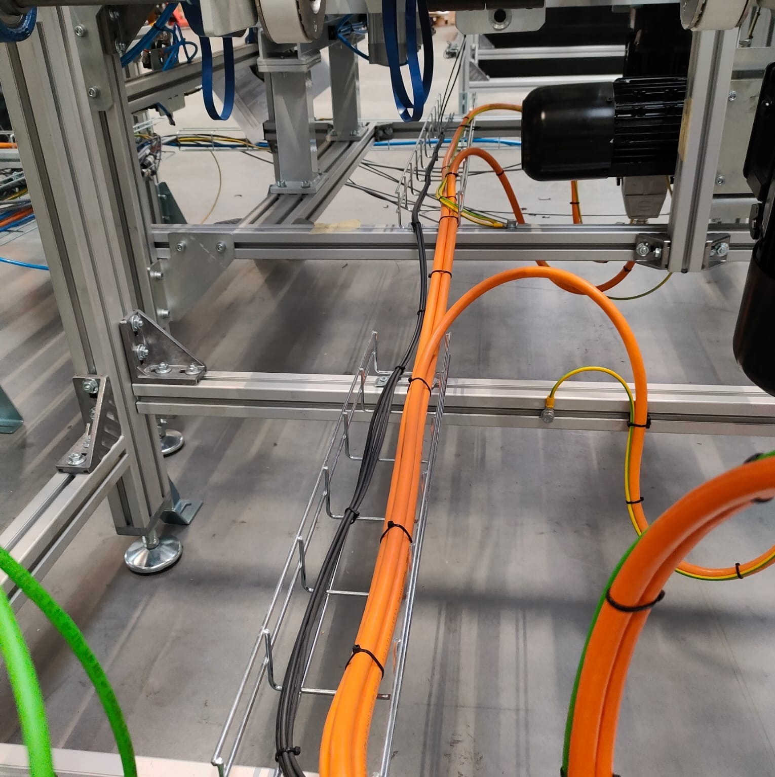 Pomarańczowe i czarne przewody elektryczne ułożone w metalowej korycie kablowej, zamocowane do aluminiowej konstrukcji z silnikami elektrycznymi w tle.