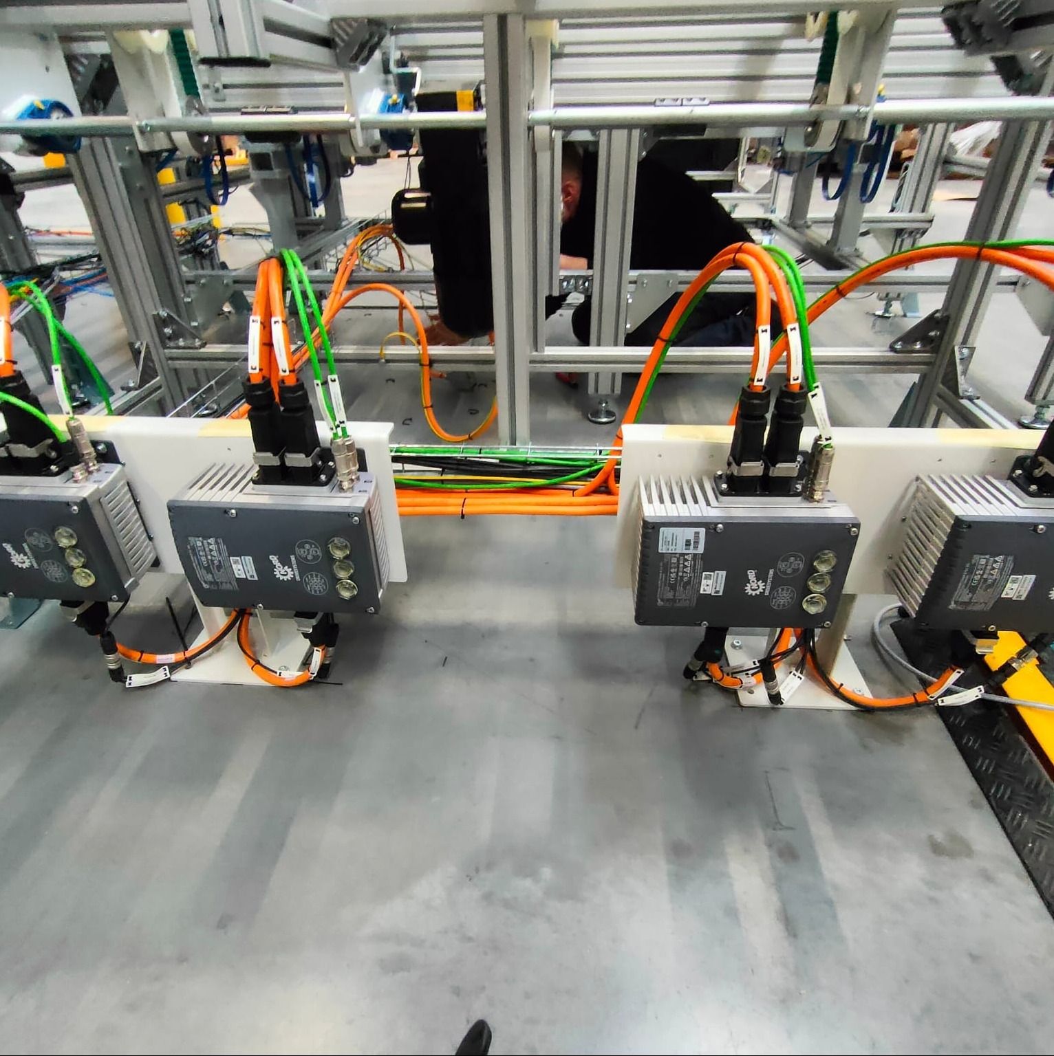 Szeregowo zamontowane moduły sterujące z okablowaniem pomarańczowo-zielonym na konstrukcji aluminiowej; widoczny fragment osoby pracującej nad instalacją w tle.