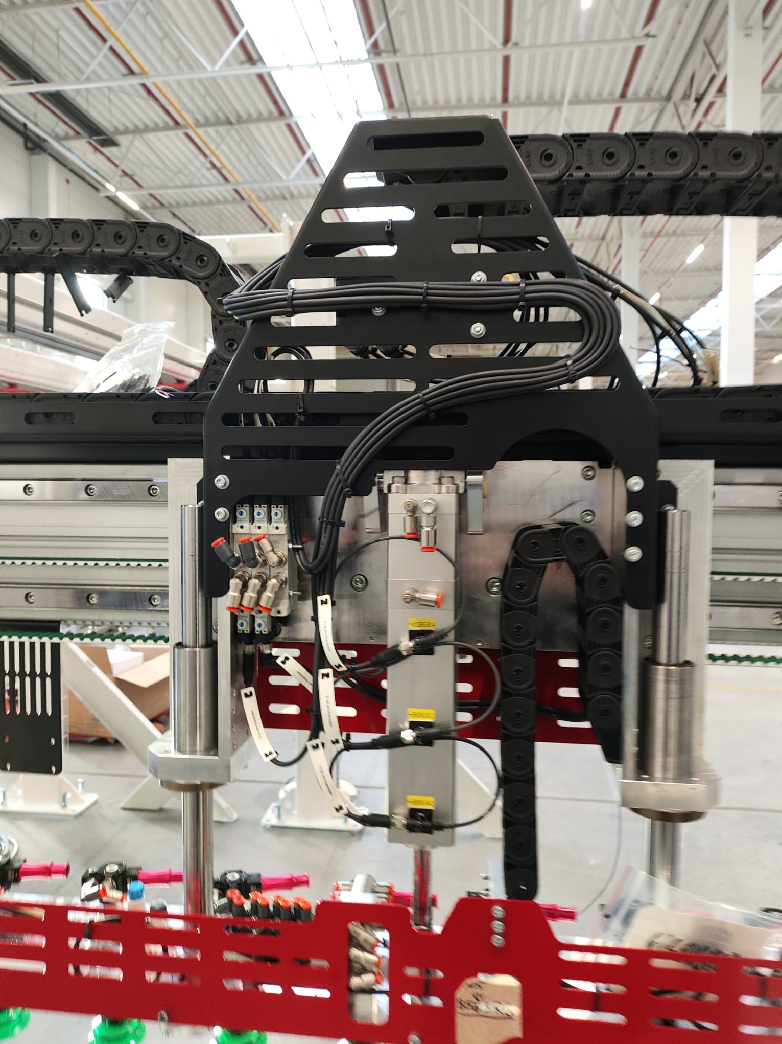 Szczegółowy widok fragmentu linii produkcyjnej z przewodami, czujnikami i elementami pneumatycznymi, umieszczonego w hali przemysłowej z widoczną konstrukcją dachu.