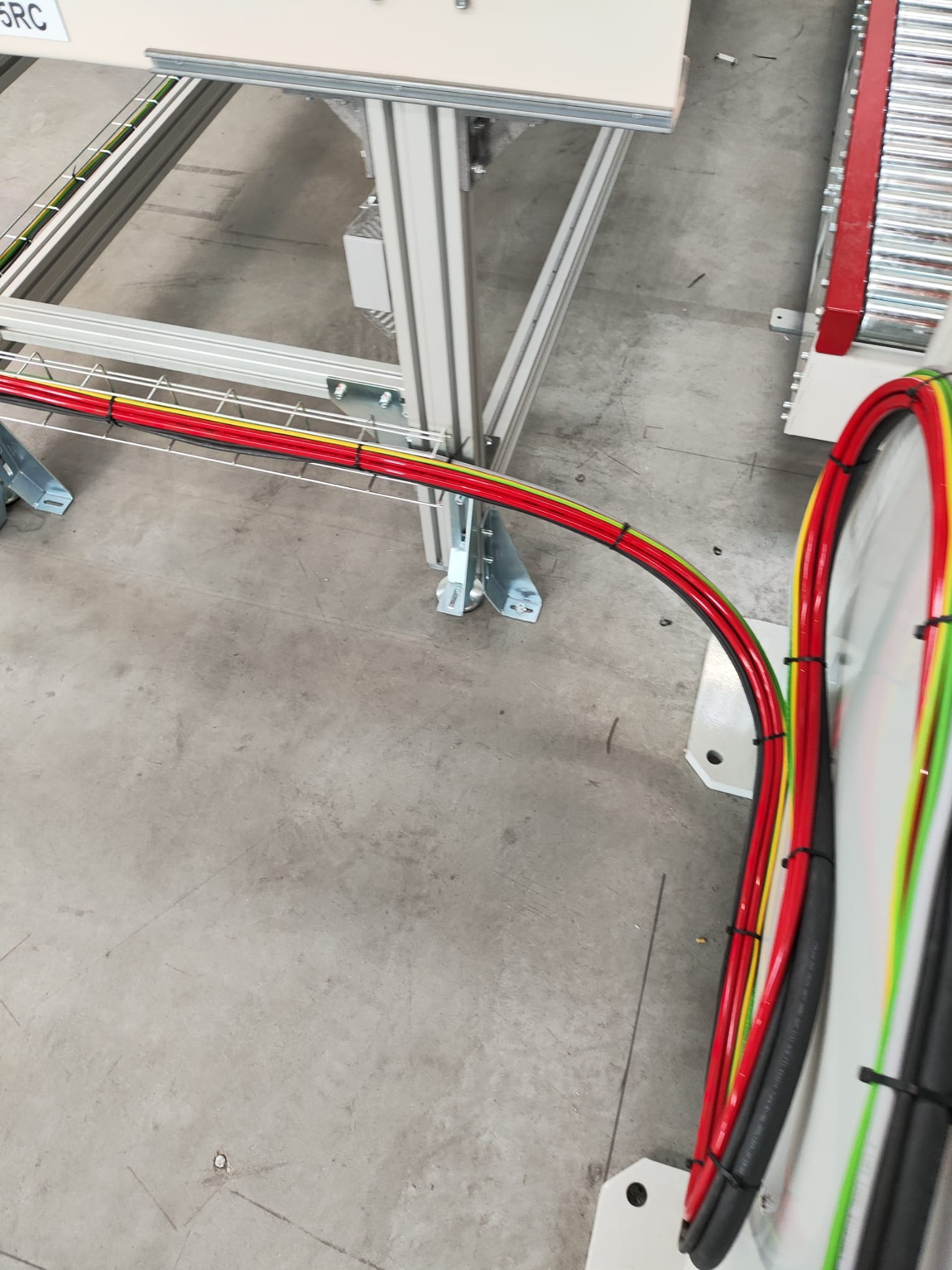 Kable elektryczne w metalowej korycie kablowej, poprowadzone wzdłuż ramy urządzenia przemysłowego, zabezpieczone opaskami zaciskowymi, widoczne kolory izolacji: czerwony, żółty, zielony.