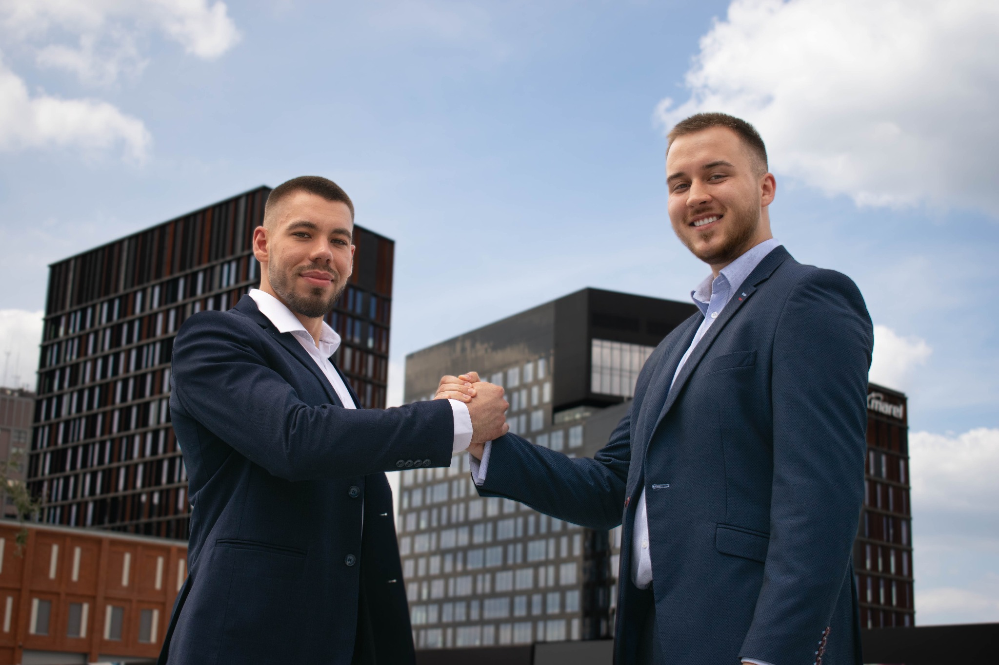 Dwóch uśmiechniętych mężczyzn w garniturach ściska sobie dłonie na tle nowoczesnych budynków biurowych i błękitnego nieba z chmurami.
