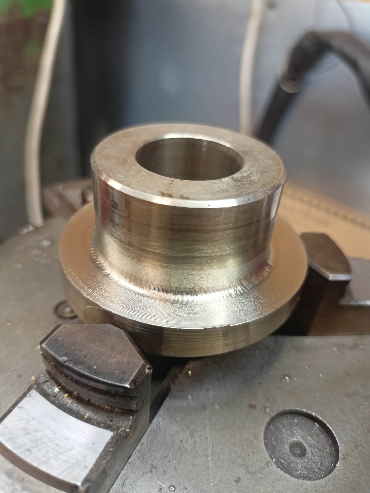 Metalowy element z otworem, obrobiony i zamocowany w uchwycie tokarskim. Widoczne ślady obróbki i spawania.