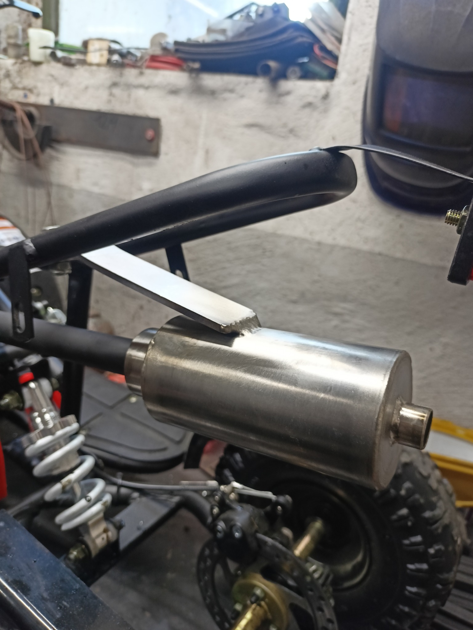 Spawany, nierdzewny tłumik zamontowany na pojeździe typu ATV, widoczny fragment spawu, tło warsztatowe z maską spawalniczą.