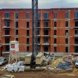 DenGlass - Budynek w trakcie budowy z zamontowanymi oknami, widoczny żuraw, materiały budowlane i samochody na placu budowy.