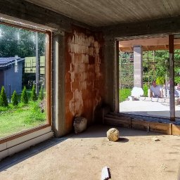 DenGlass - Surowy stan wnętrza z dużymi otworami okiennymi, widok na ogród z panelami słonecznymi i pracującego mężczyznę na zewnątrz, mur z cegły dziurawki.