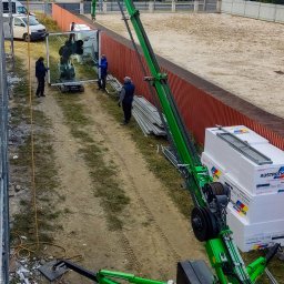 DenGlass - Montaż okna z użyciem specjalistycznego dźwigu ssącego; trzech pracowników w odzieży roboczej nadzoruje proces, w tle widoczne budynki mieszkalne.