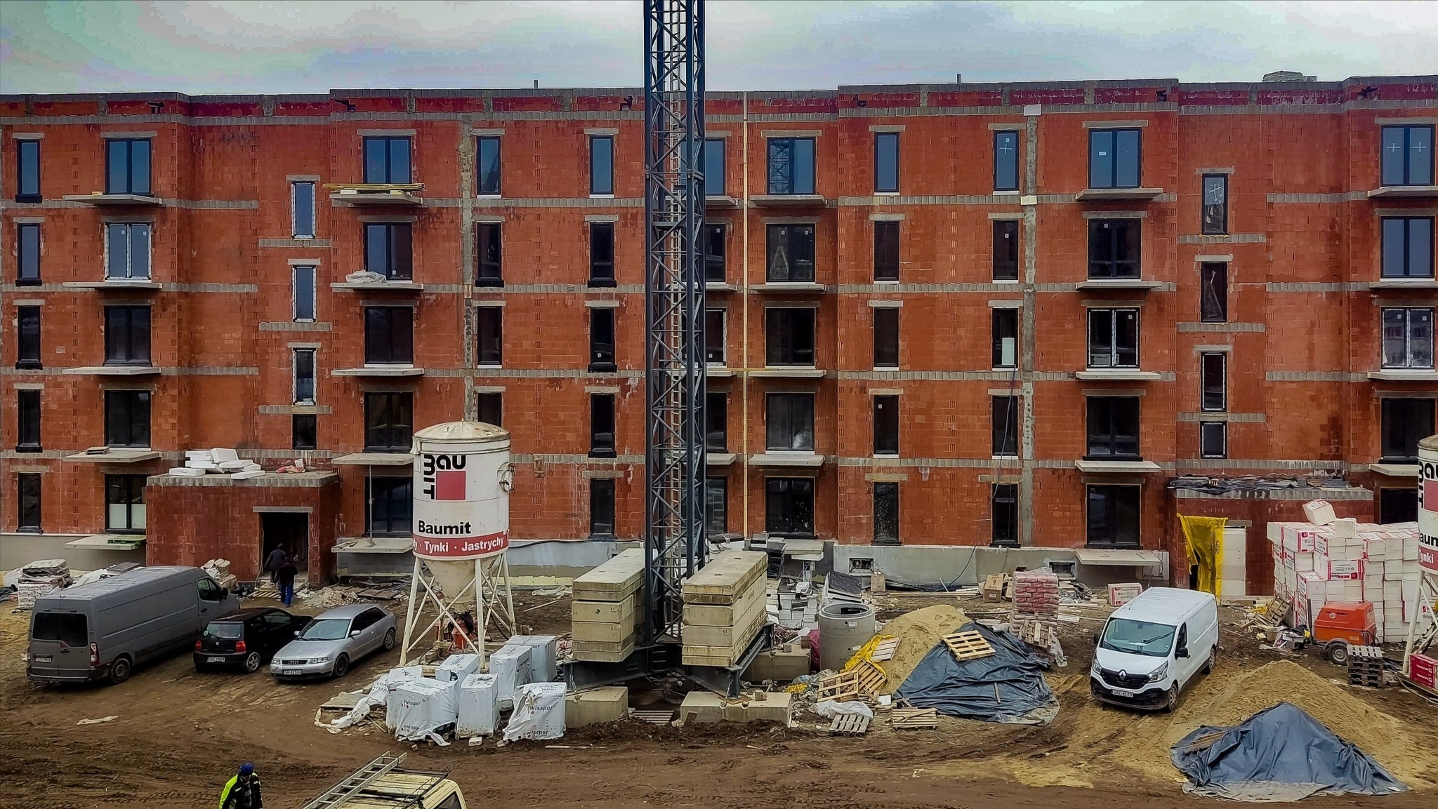 Budynek w trakcie budowy z zamontowanymi oknami, widoczny żuraw, materiały budowlane i samochody na placu budowy.