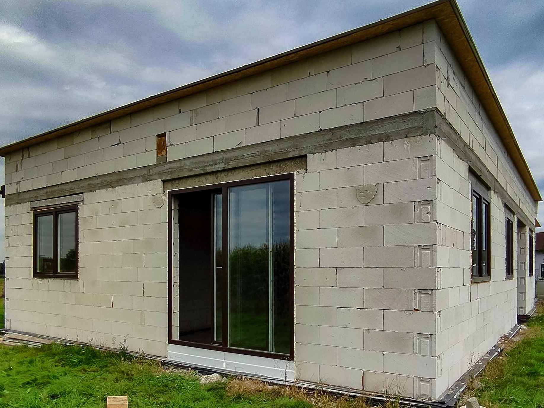 Surowy stan budynku z betonu komórkowego z zamontowanymi oknami PCV i przesuwnymi drzwiami tarasowymi, widok z zewnątrz na tle pochmurnego nieba.