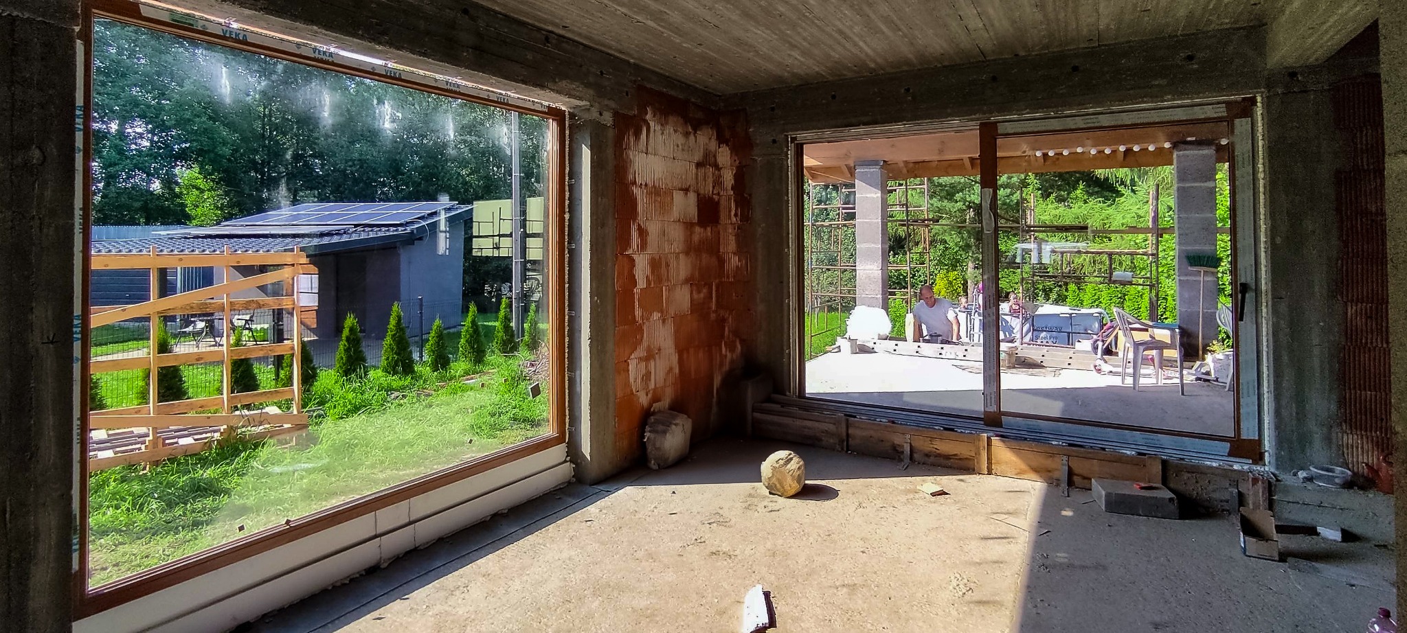 Surowy stan wnętrza z dużymi otworami okiennymi, widok na ogród z panelami słonecznymi i pracującego mężczyznę na zewnątrz, mur z cegły dziurawki.