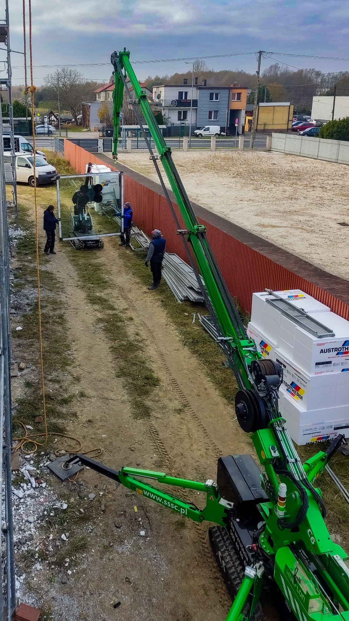 Montaż okna z użyciem specjalistycznego dźwigu ssącego; trzech pracowników w odzieży roboczej nadzoruje proces, w tle widoczne budynki mieszkalne.