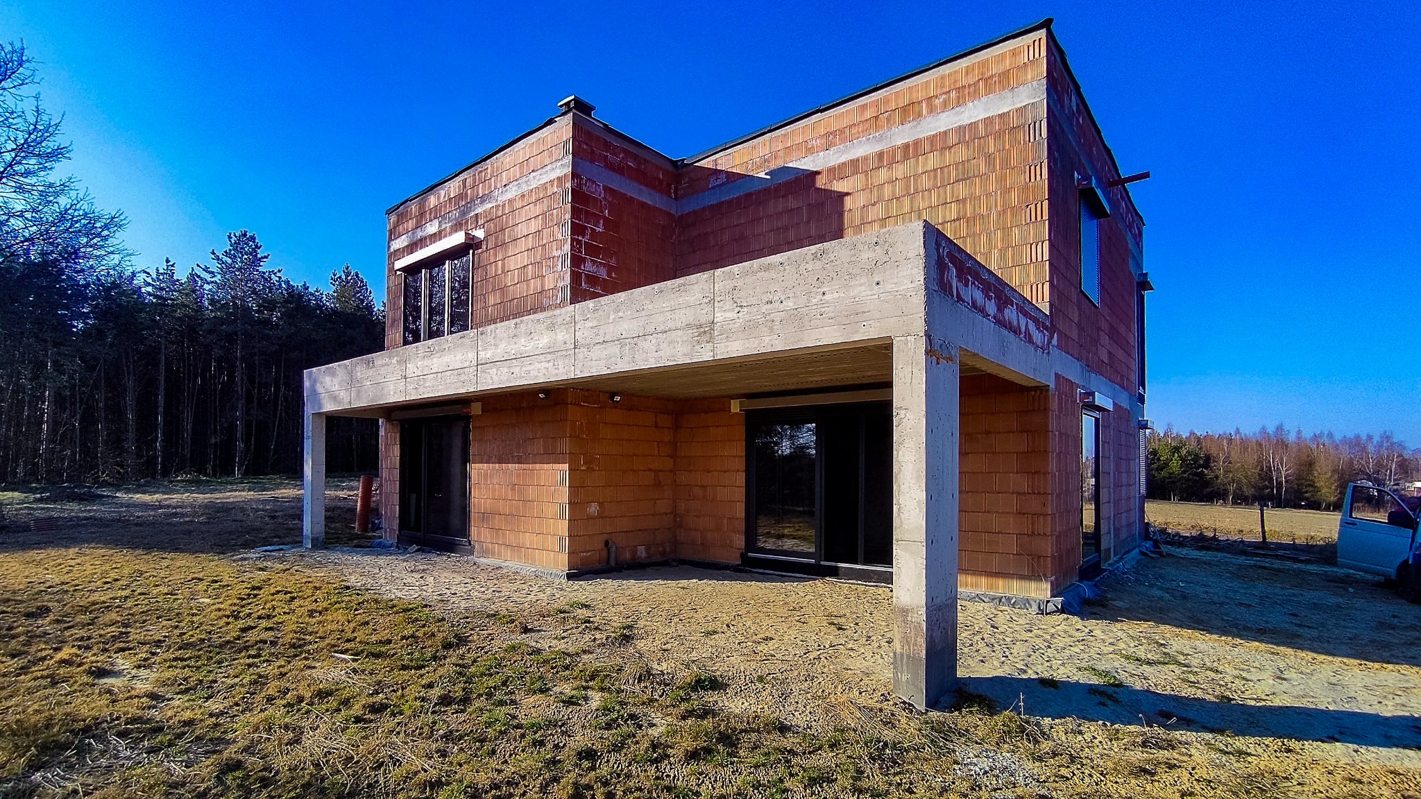 Dom w trakcie budowy, widoczne okna z ciemnymi ramami, ściany z czerwonej cegły, betonowy zadaszony taras, surowy stan, błękitne niebo, las w tle.