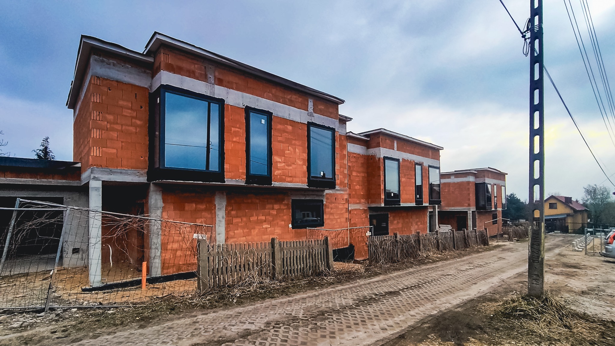 Surowy stan budowy bliźniaków z zamontowanymi oknami PCV w czarnych ramach, widoczne bloczki ceramiczne i betonowe nadproża, teren budowy ogrodzony, słup energetyczny po prawej.