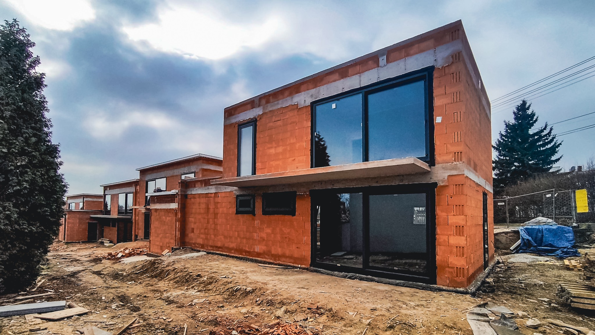 Surowy stan budowy szeregu domów jednorodzinnych z dużymi oknami z czarnymi ramami, widok na plac budowy z porozrzucanymi cegłami i narzędziami, pochmurne niebo w tle.