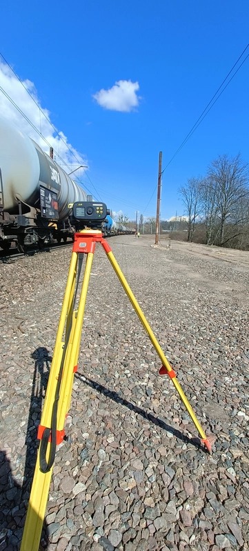 Żółty niwelator na trójnogu ustawiony na żwirowym podłożu, w tle pociąg towarowy i błękitne niebo z pojedynczą chmurą.