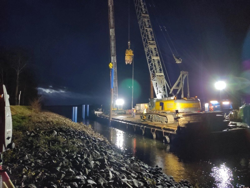 Nocne prace geodezyjne przy budowie mostu: dźwig na barce wbija pale w rzece, oświetlone reflektorami, widoczne brzegi rzeki z kamieniami.
