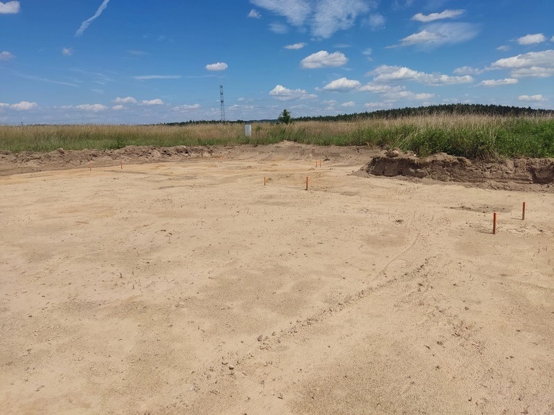 Wytyczanie fundamentów pod budowę w Szczecinie, widoczne pomarańczowe paliki geodezyjne na piaszczystym terenie.