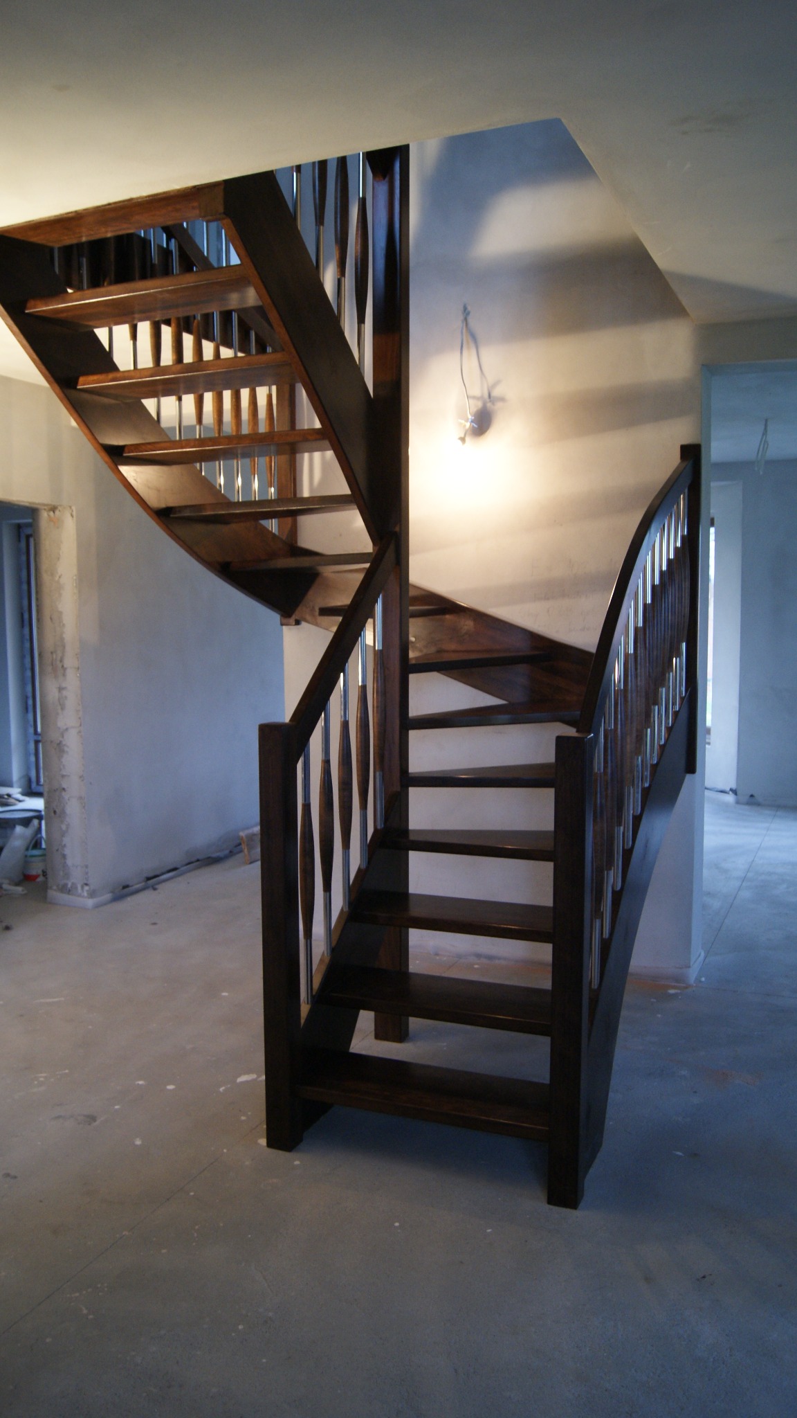 Ciemnobrązowe schody z metalowymi tralkami, zakręcające ku górze, w surowym wnętrzu w trakcie wykańczania.