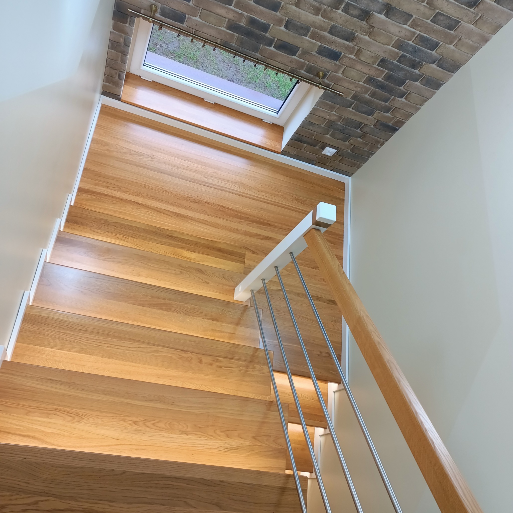 Nowoczesne schody drewniane z jasnego drewna z metalową balustradą, oświetleniem LED pod stopniami i oknem dachowym z widokiem na zieleń.