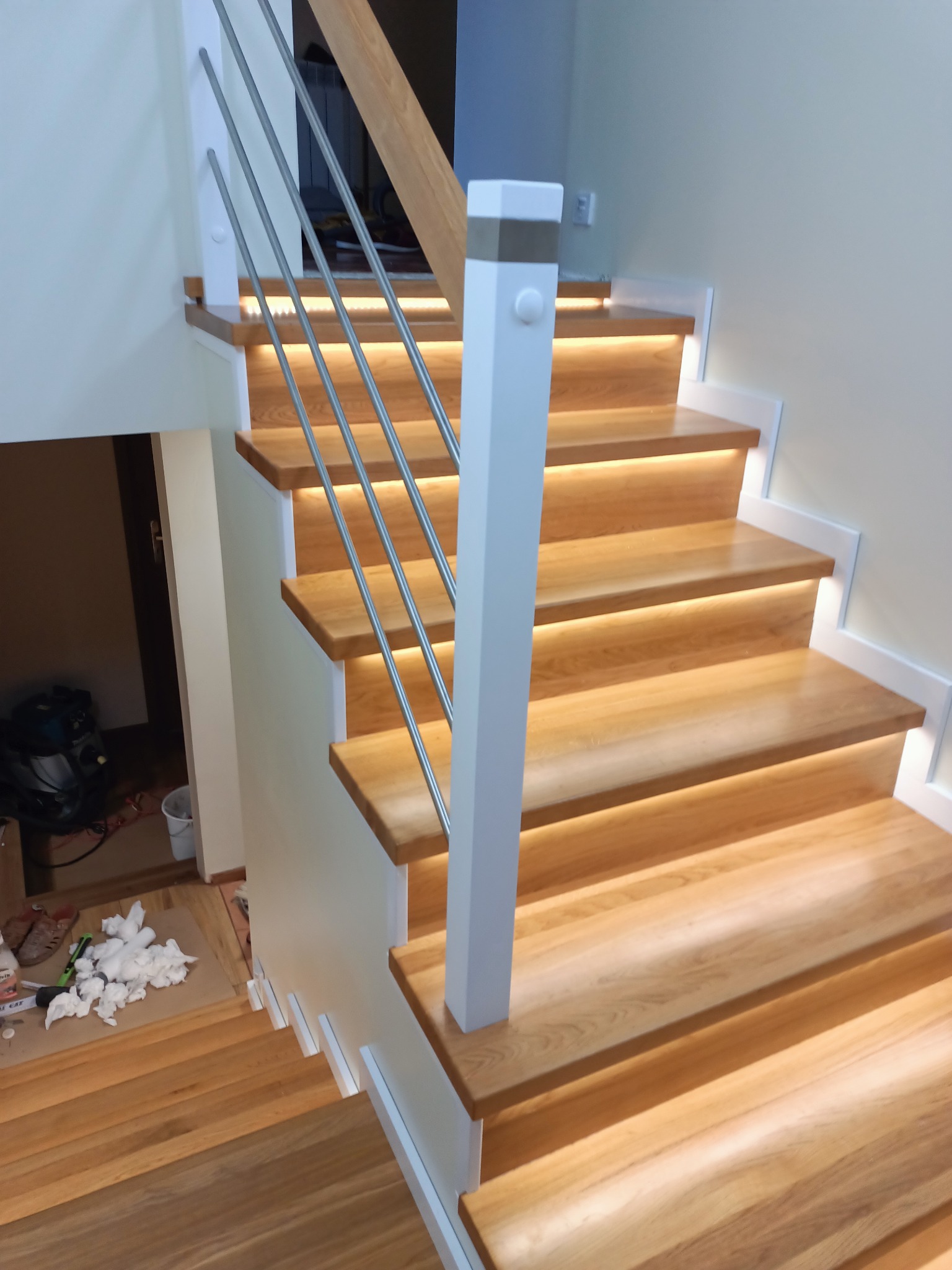 Jasne, drewniane schody z podświetleniem LED pod każdym stopniem, białe balustrady i stalowe pręty, widok z dołu.