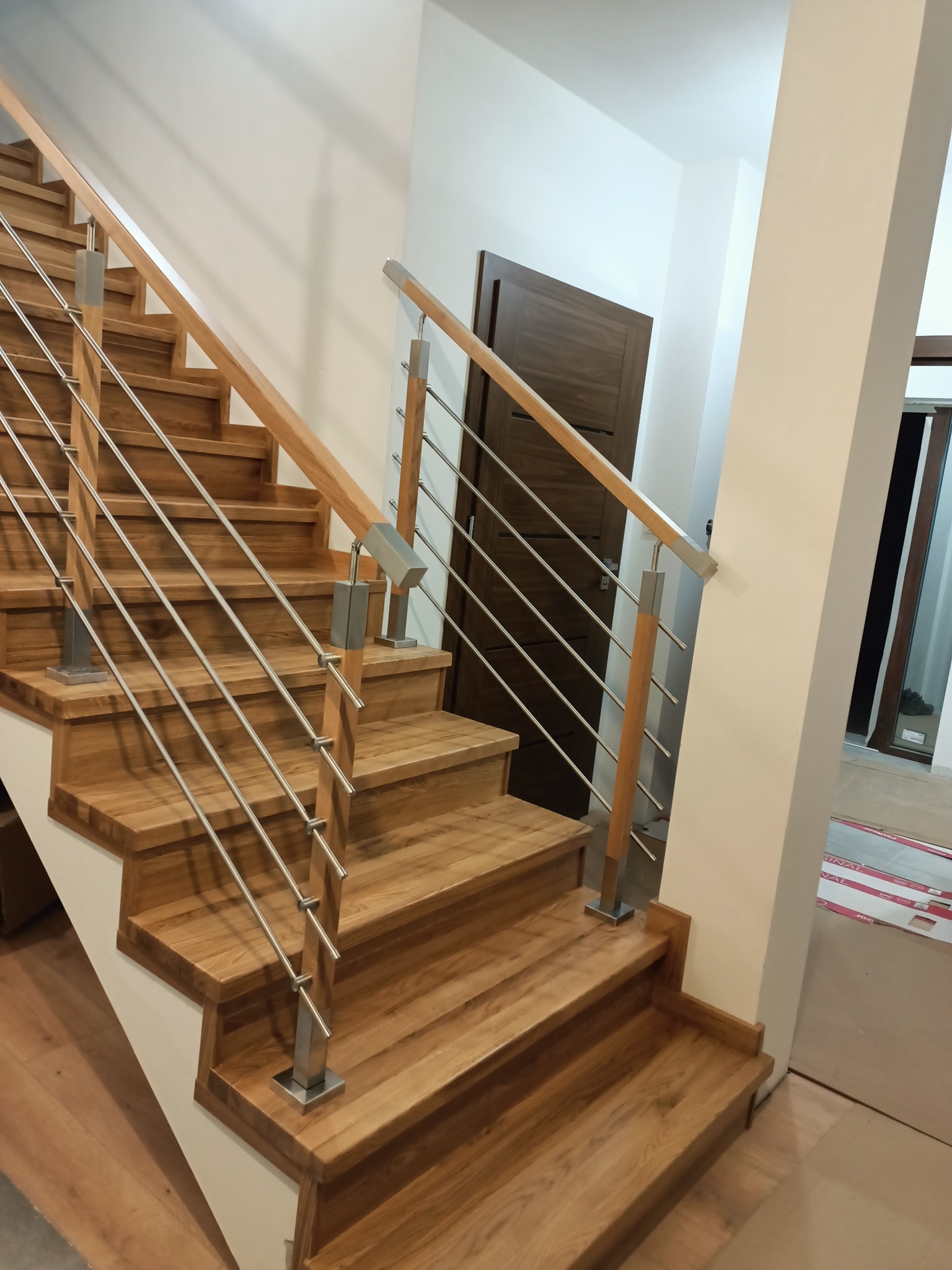 Jasne, drewniane stopnie schodów z metalową balustradą w nowoczesnym wnętrzu, widoczny fragment drzwi w tle.