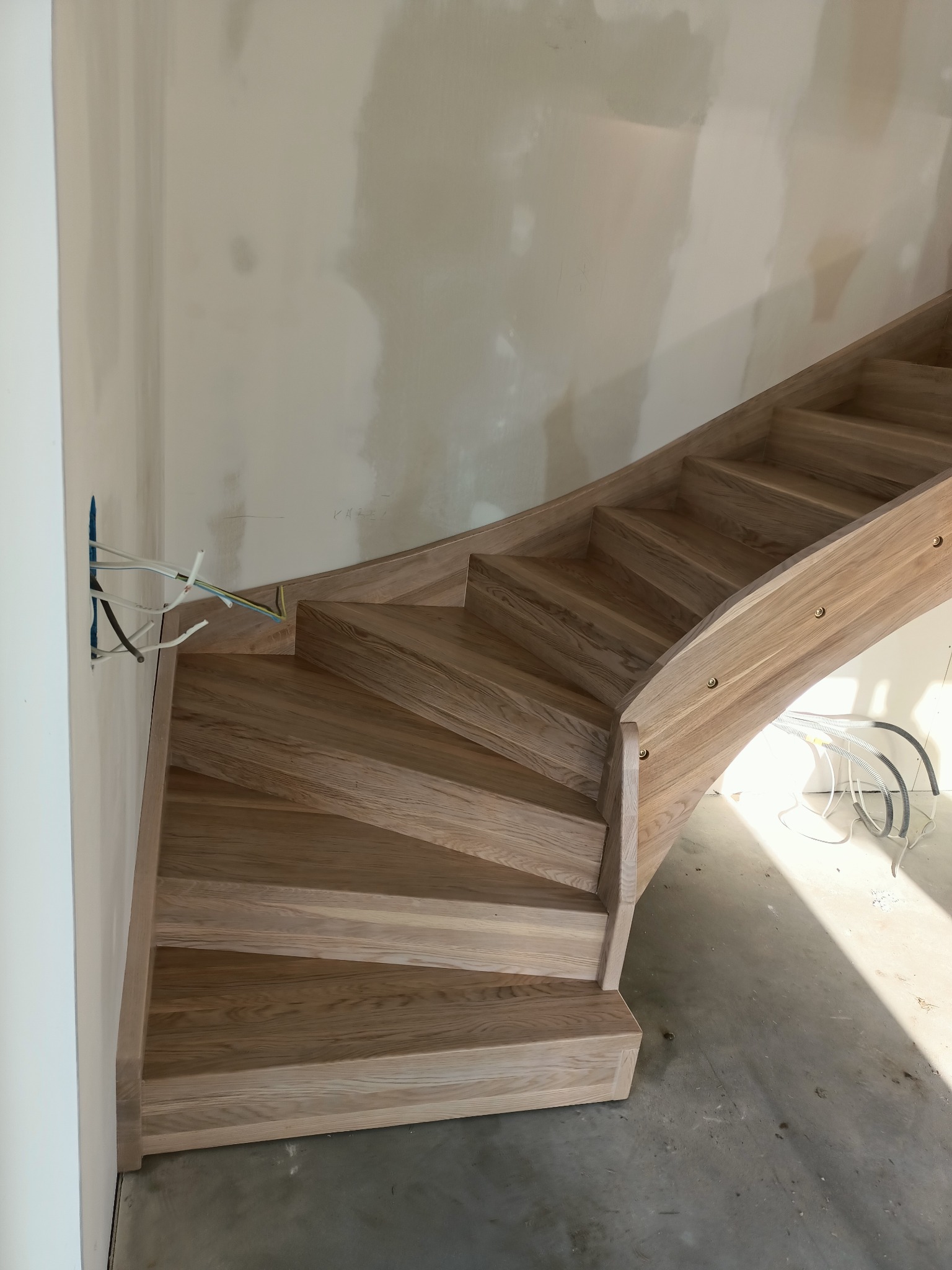 Zakręcone, jasne, dębowe schody w trakcie montażu, widoczne przewody elektryczne wystające ze ściany i surowy beton na podłodze.