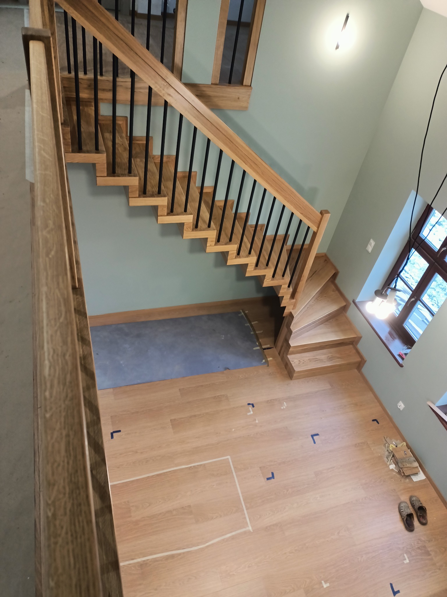 Wysoki kąt widzenia na jasne, drewniane schody z czarnymi balustradami w nowoczesnym domu, z widocznymi śladami po taśmie malarskiej na podłodze i oknem w tle.