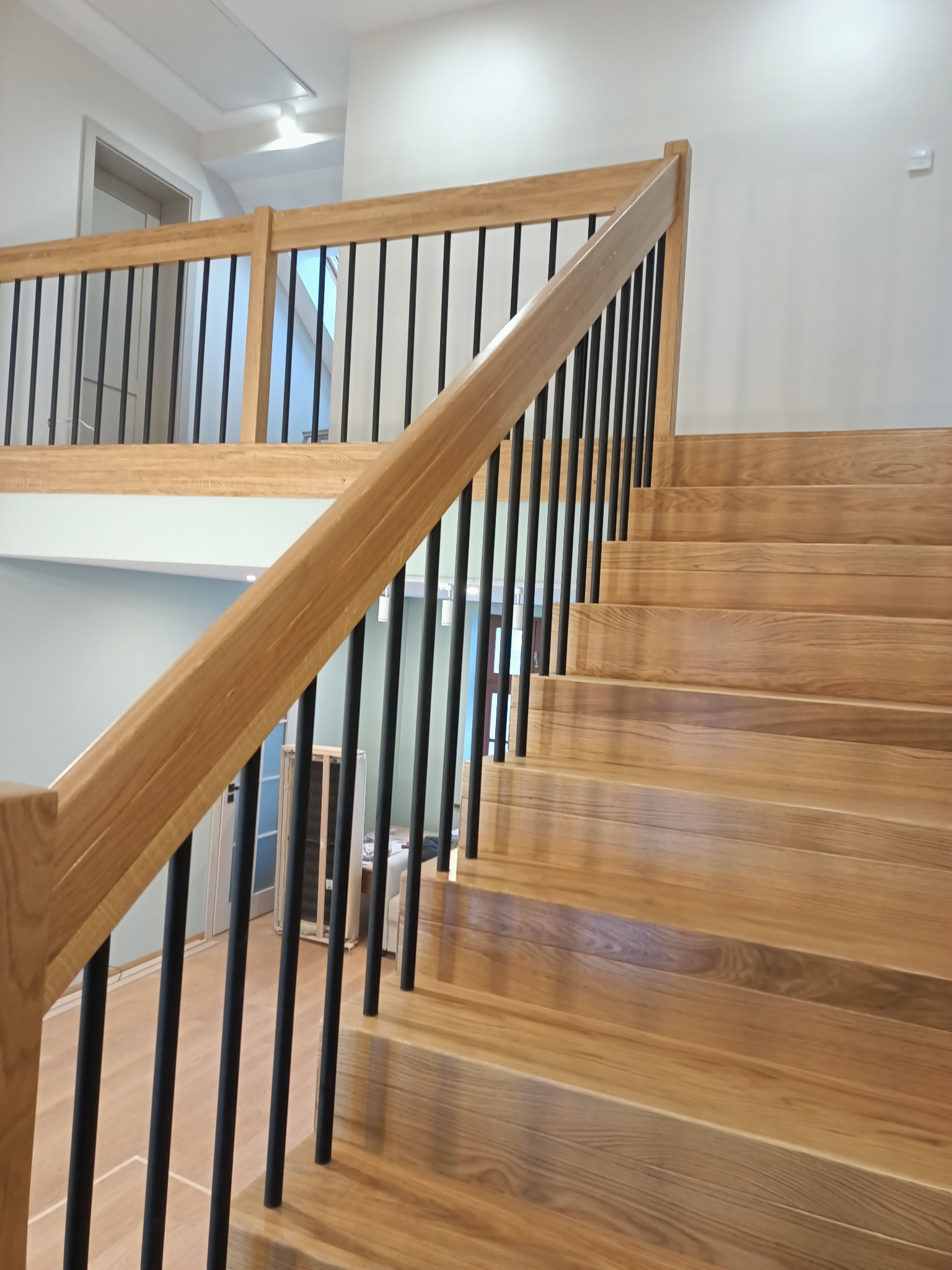 Jasne drewniane schody z czarnymi, metalowymi balustradami w nowoczesnym wnętrzu domu, widok z dołu na górę biegu schodów.