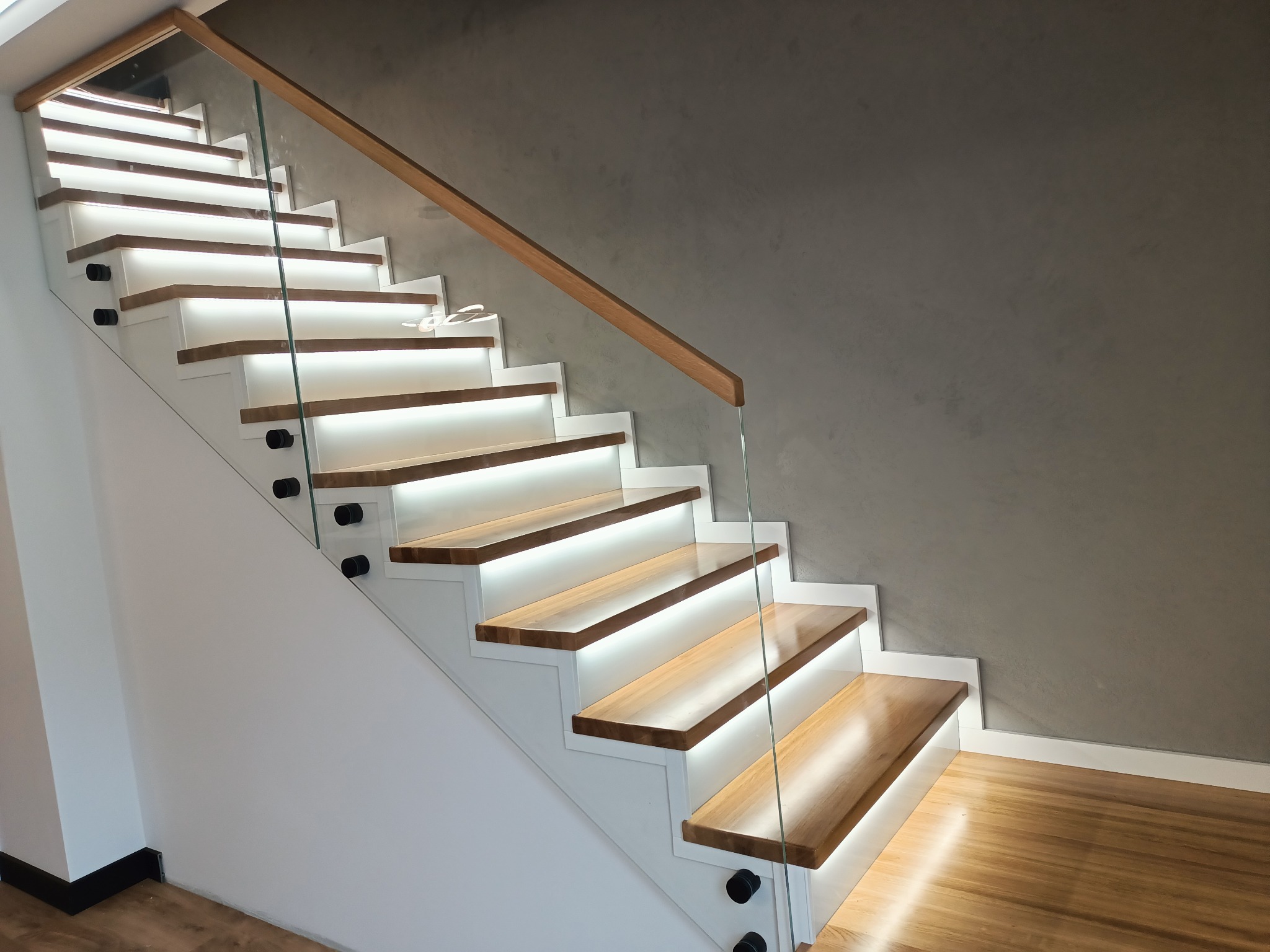Nowoczesne schody z podświetlanymi stopniami i szklaną balustradą, widok z dołu, drewniane stopnie na białej konstrukcji.