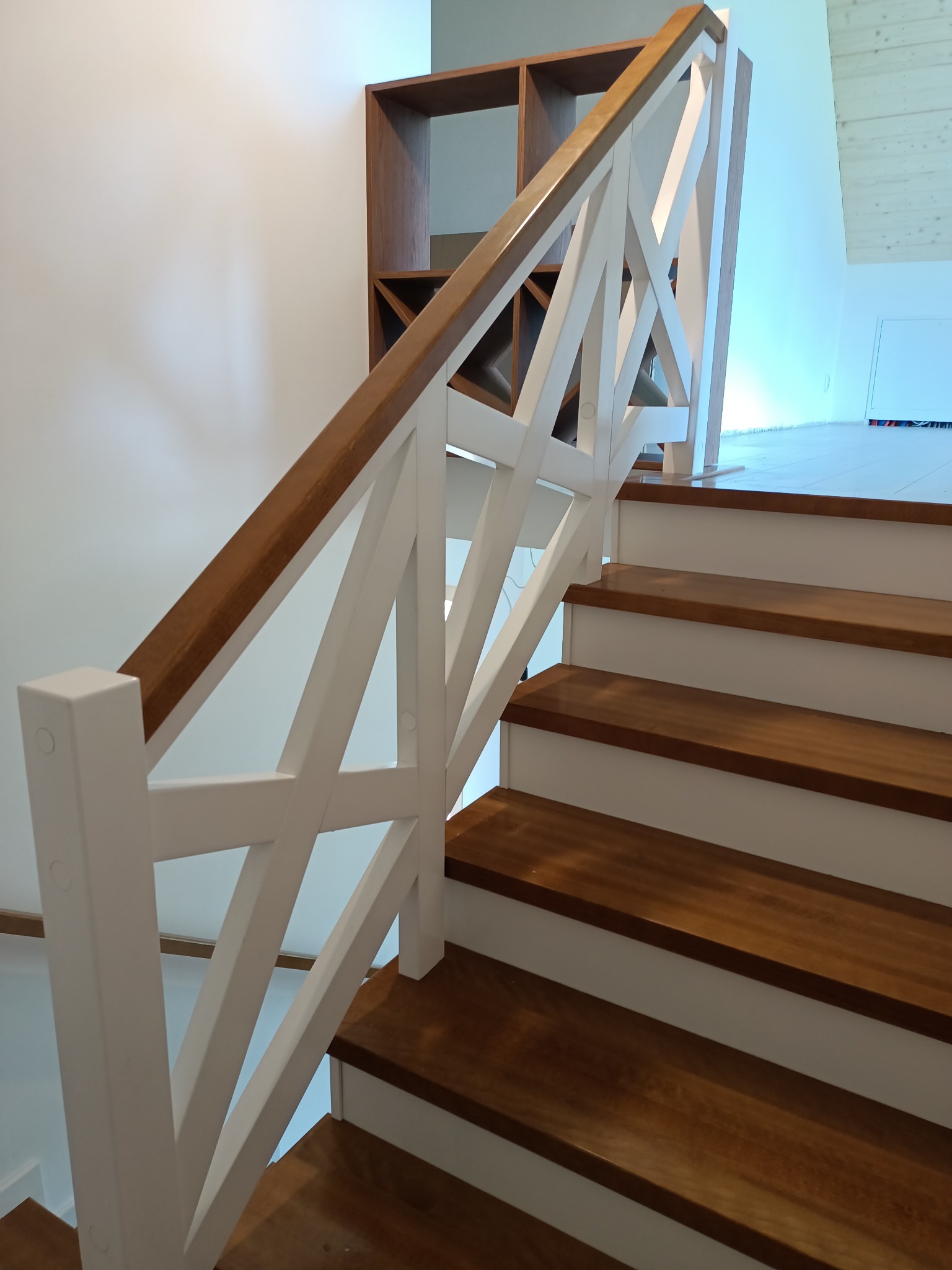 Drewniane schody z białymi podstopnicami i balustradą w stylu krzyżowym, z brązowym drewnianym pochwytem, widok z dołu na pierwsze stopnie, w tle regał.
