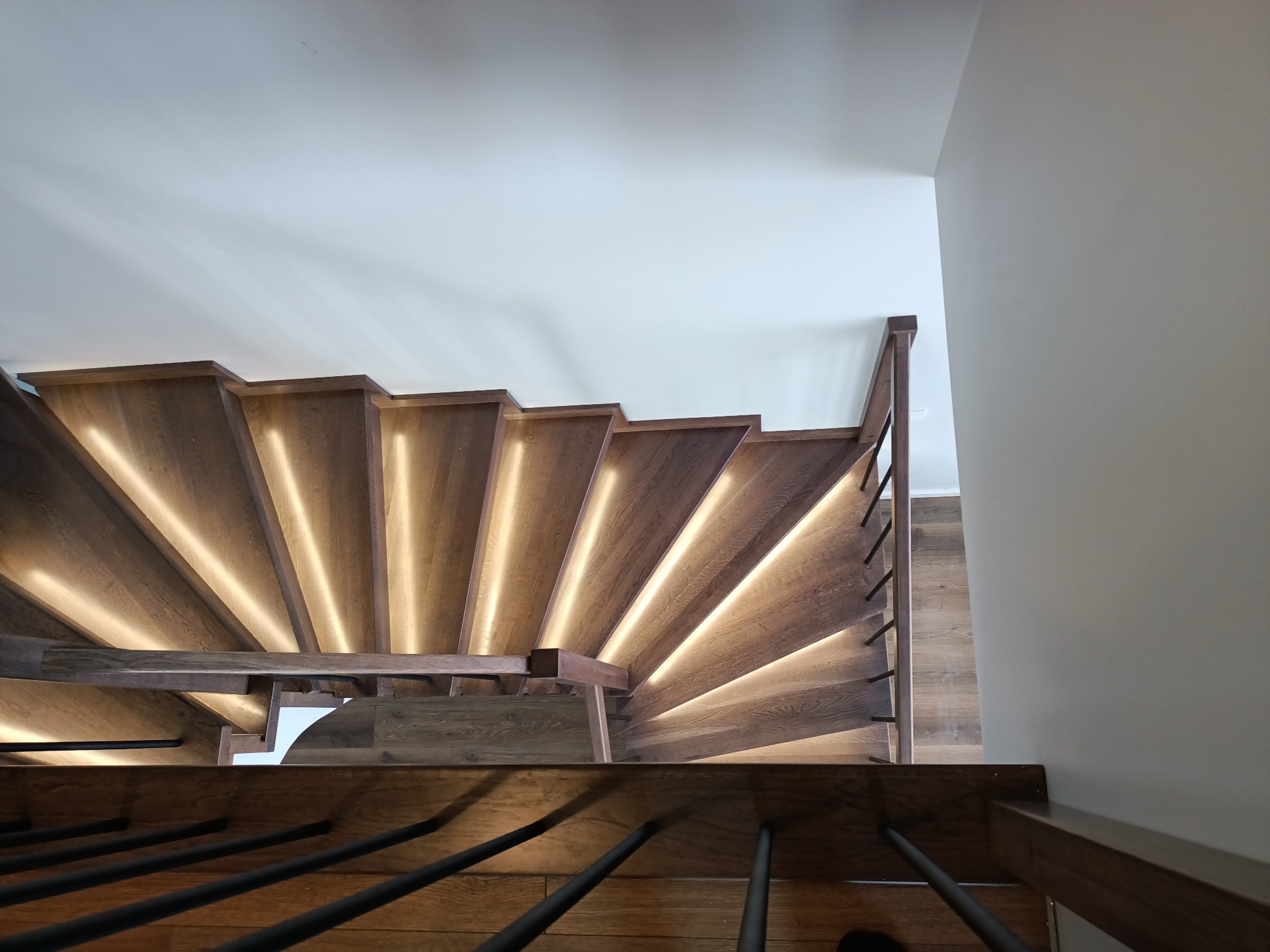 Spiralne schody z drewna z podświetleniem LED na każdym stopniu, widok z góry, czarne metalowe balustrady, jasne ściany.