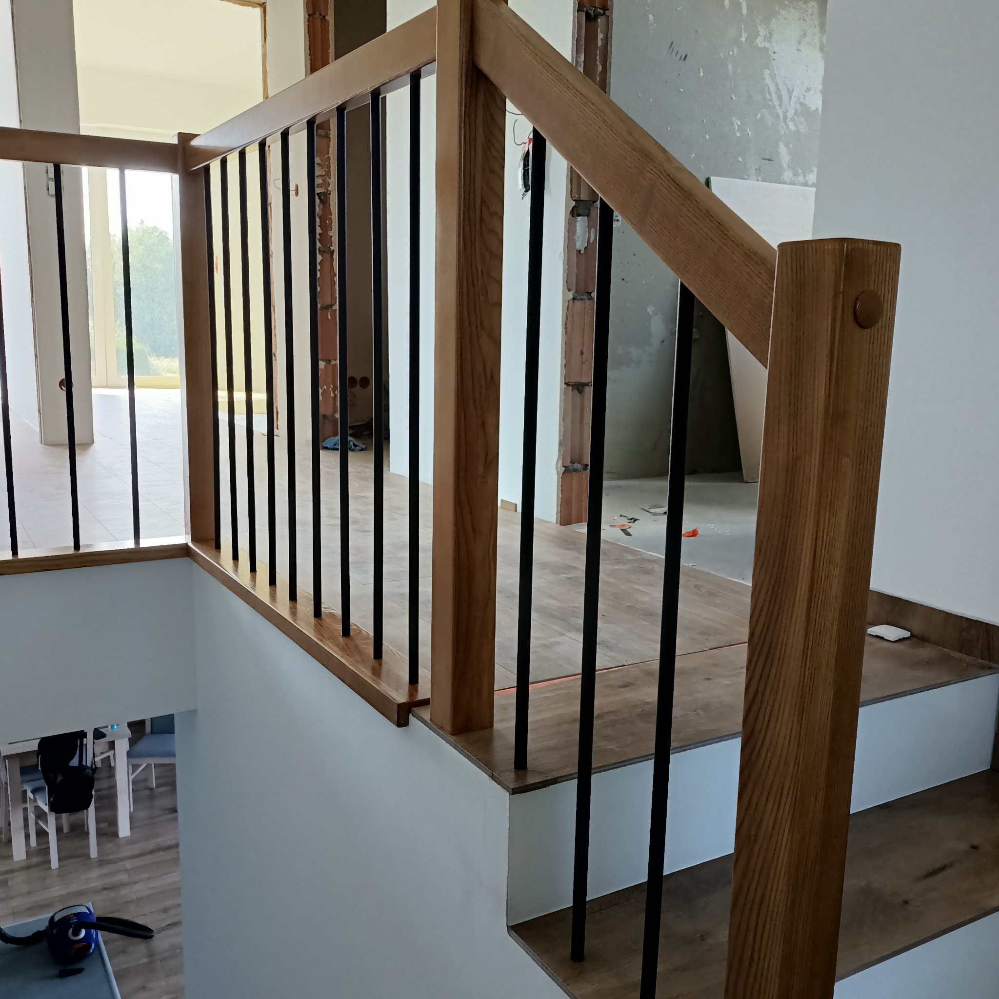 Fragment balustrady z jasnego drewna z czarnymi, pionowymi elementami na schodach wykończonych panelami imitującymi drewno. W tle widoczne surowe ściany w trakcie remontu i pomieszczenie z białymi...