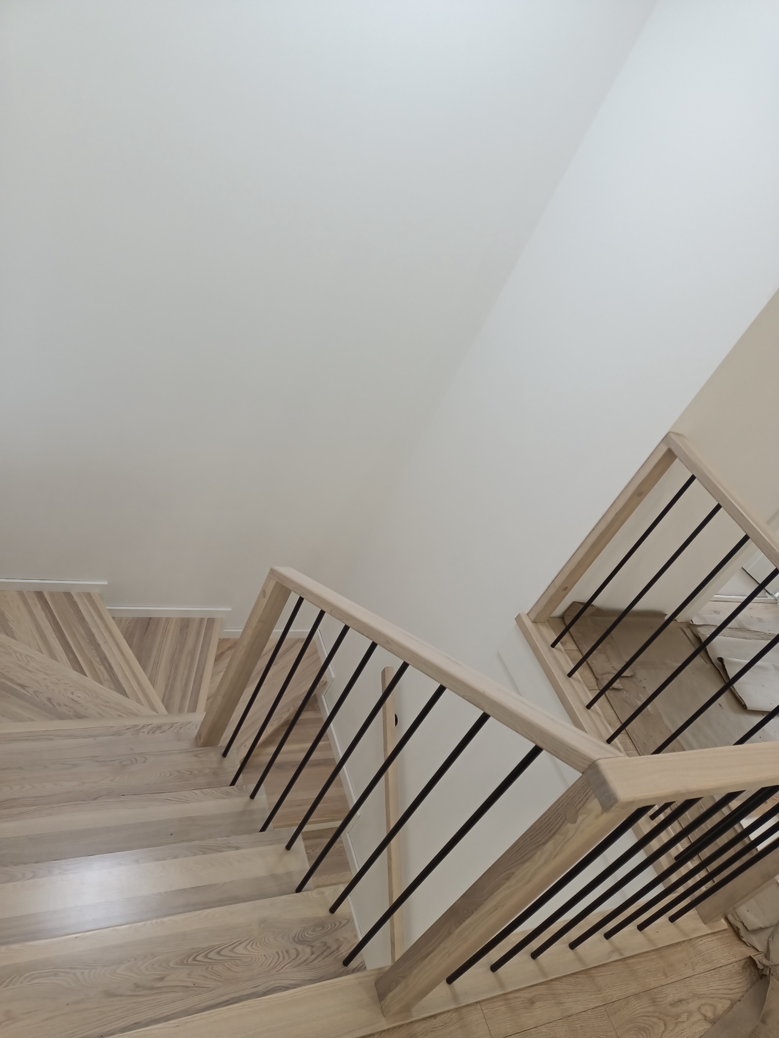 Widok z góry na jasne, drewniane schody z czarnymi, metalowymi balustradami, prowadzące w dół, z fragmentem zabezpieczonej podłogi na dole.