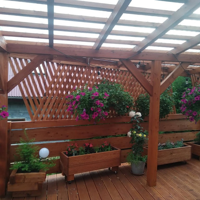 Zadaszona konstrukcja z drewna z drewnianą podłogą, ozdobiona wiszącymi i stojącymi donicami z kwiatami, oświetlona sznurem żarówek.