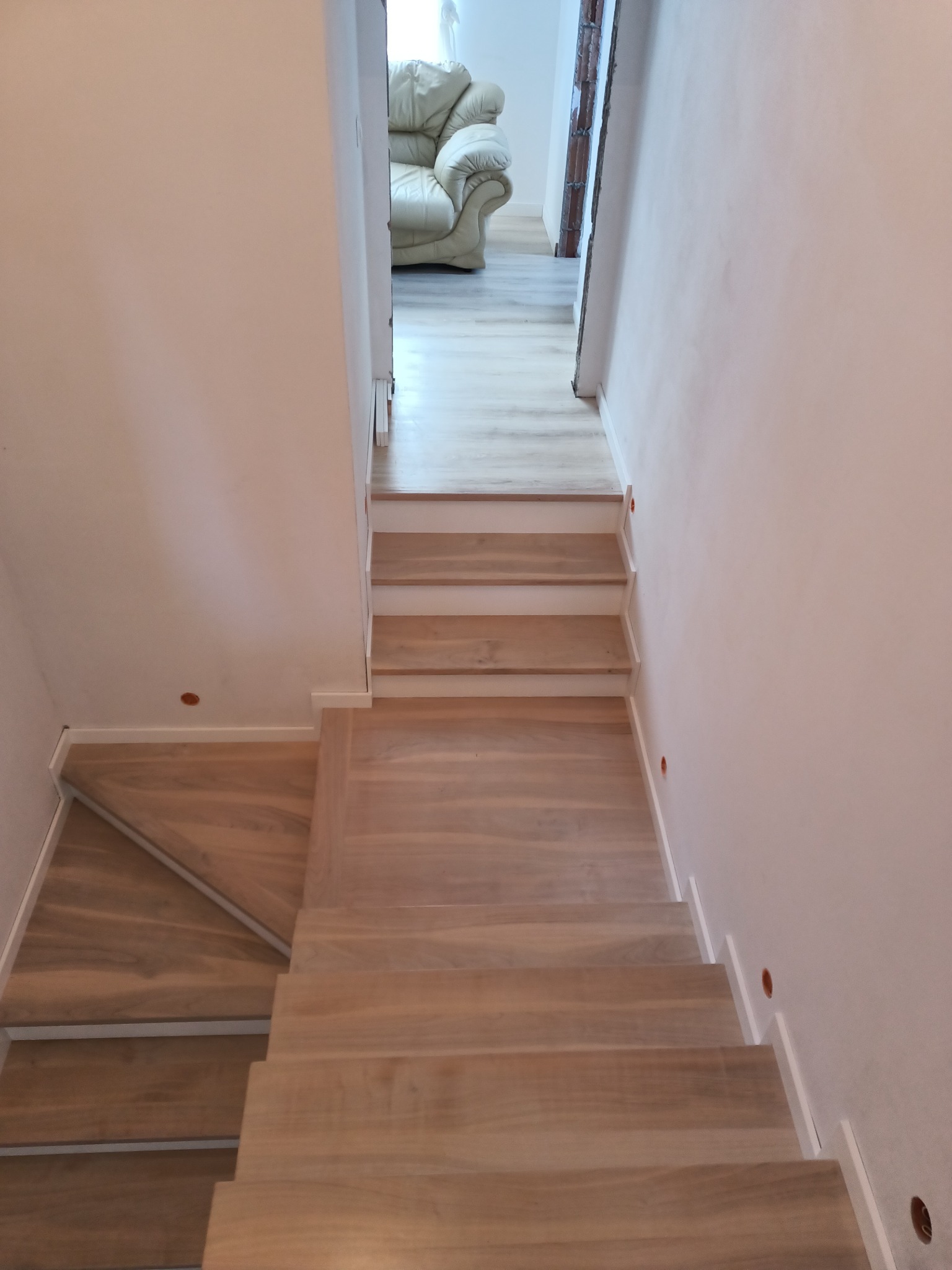 Jasne, drewniane stopnie schodów prowadzące w dół, z widocznym fragmentem białej sofy w tle.