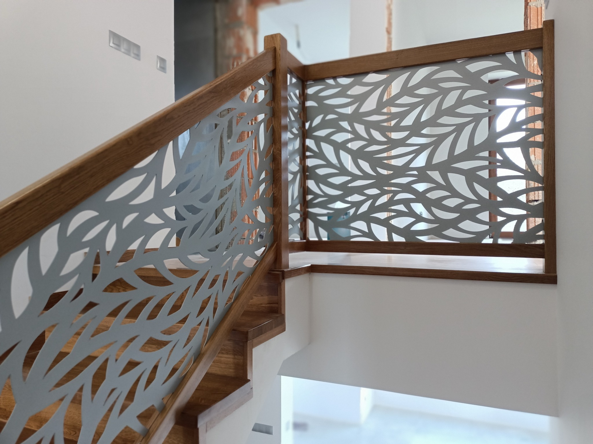 Nowoczesne schody z drewnianymi stopniami i balustradą z ażurowym, metalowym wypełnieniem o wzorze liściastym, w jasnym wnętrzu.