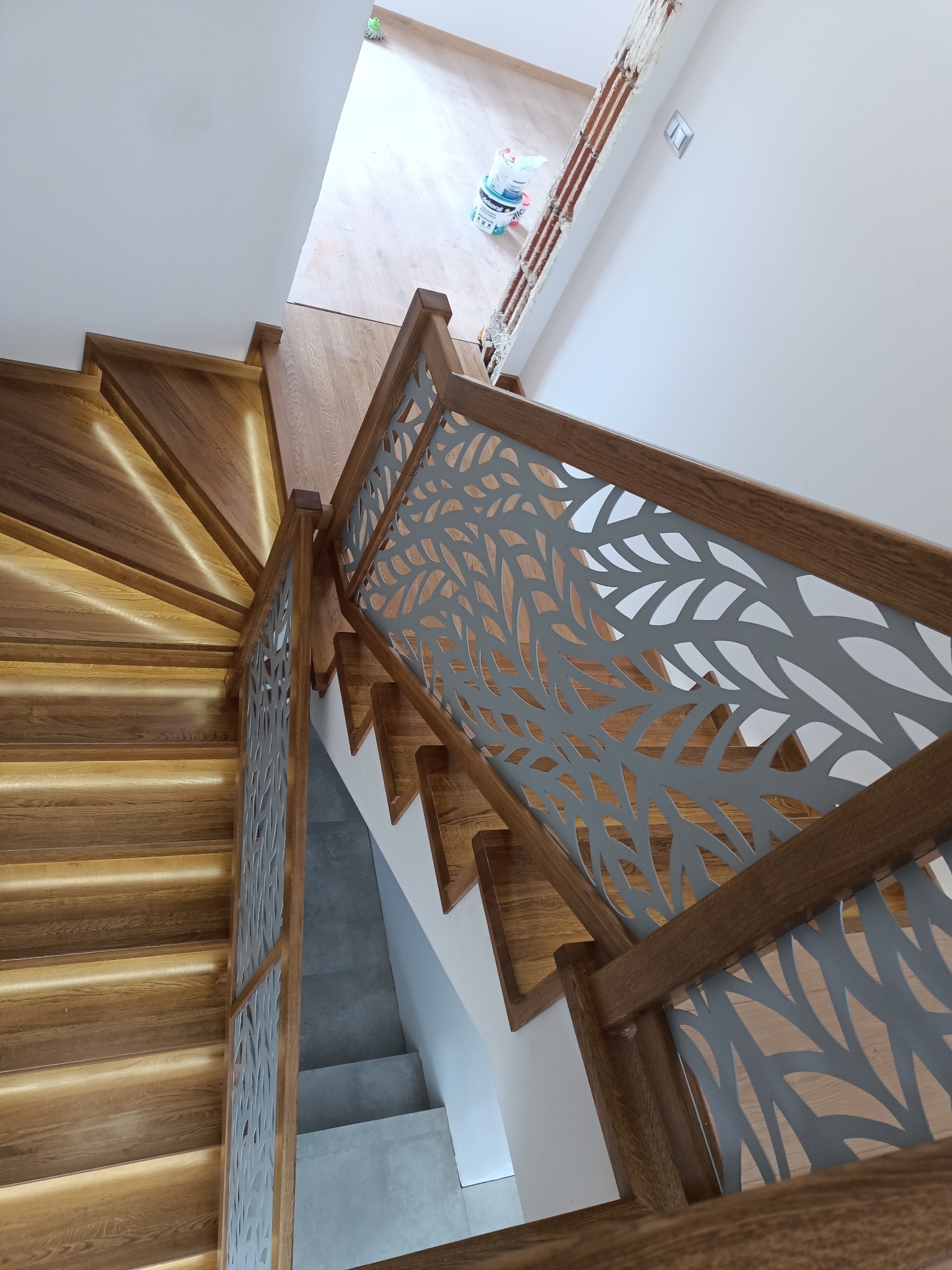 Drewniane schody ze zintegrowanym oświetleniem LED i ażurową balustradą z motywem liści, widok z góry na zakręt schodów.