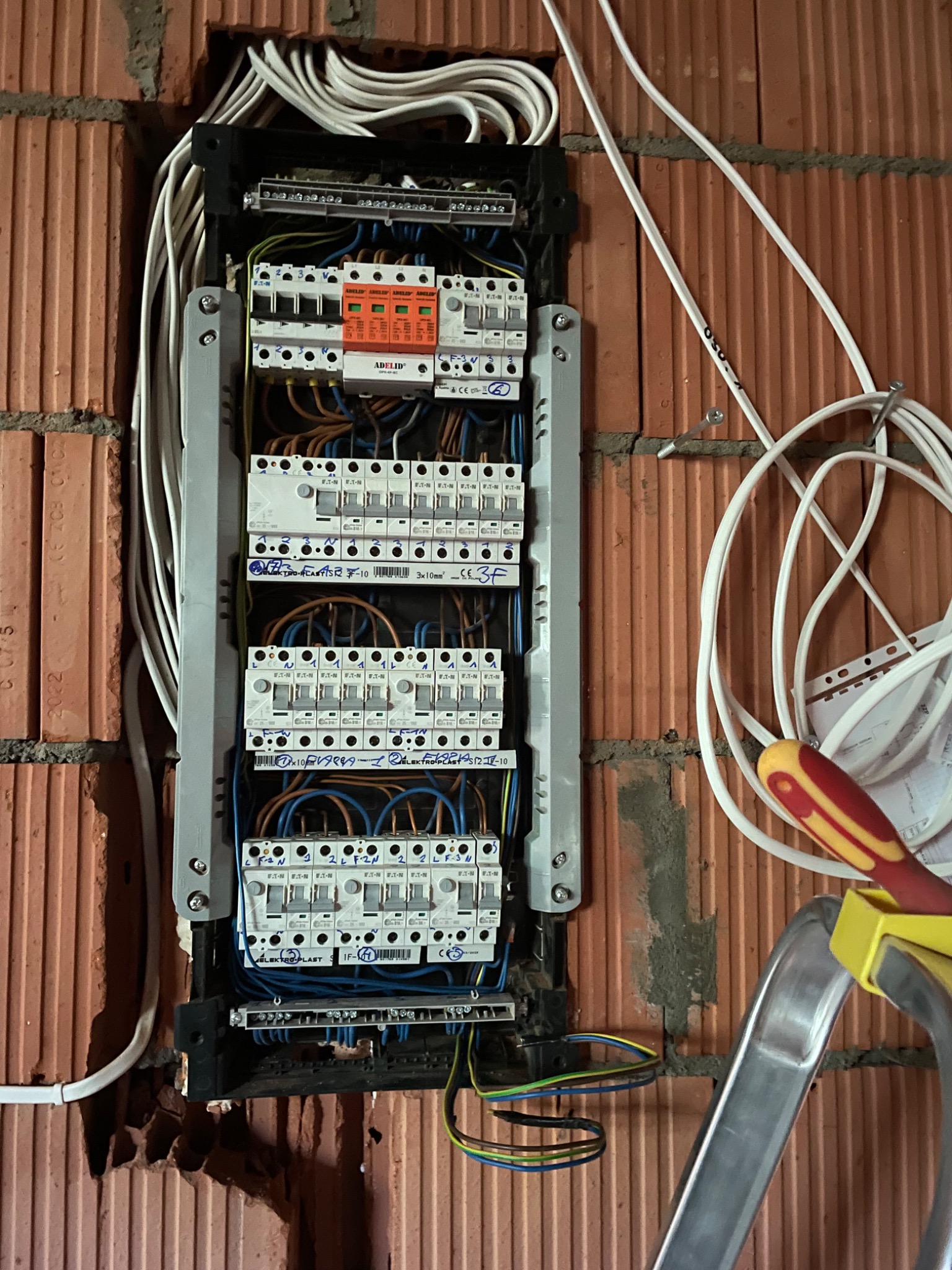 Rozdzielnica elektryczna zamontowana na ceglanej ścianie, widoczne zabezpieczenia, przewody i drabina. Instalacja w trakcie montażu.
