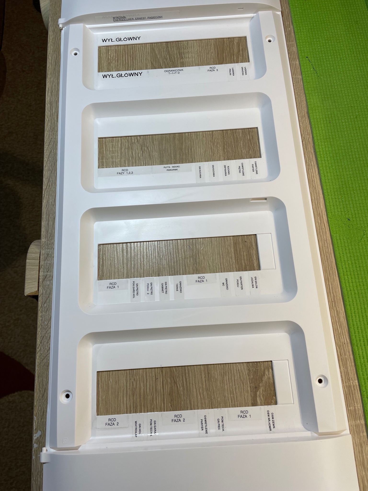 Rozdzielnica elektryczna z opisami obwodów, widok z góry. Biała obudowa z etykietami identyfikującymi fazy i pomieszczenia. Widoczne elementy montażowe.