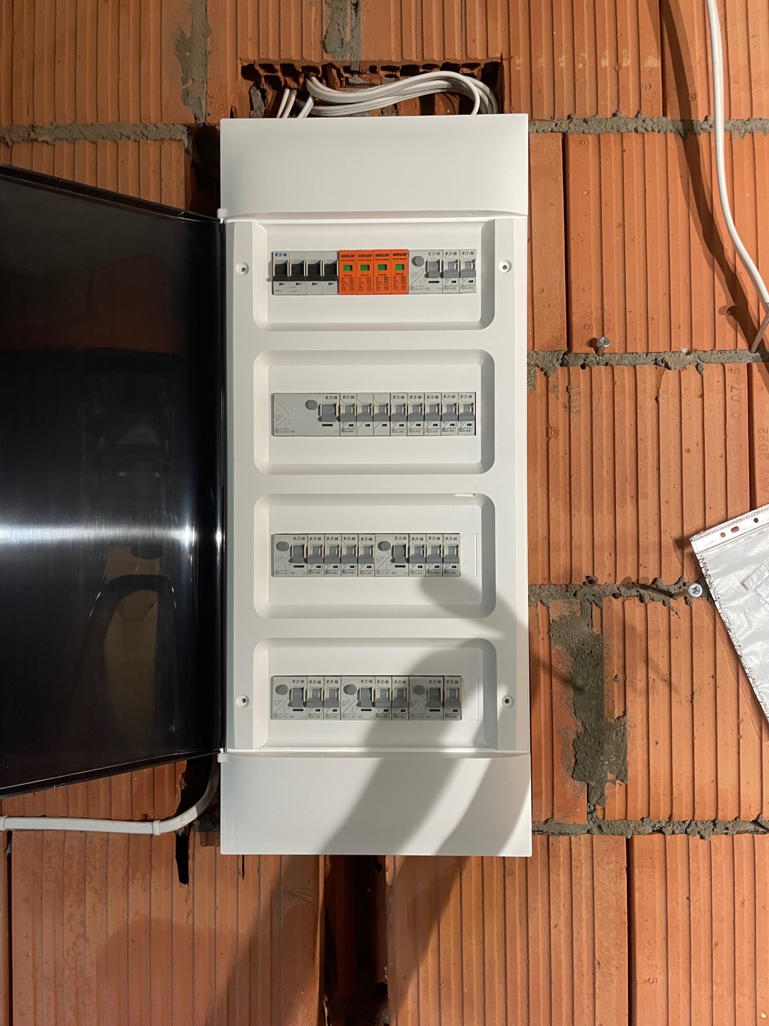 Rozdzielnica elektryczna marki Eaton z otwartymi drzwiczkami, zamontowana na ścianie z cegły dziurawki. Widoczne zabezpieczenia i przewody.