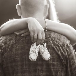 FQ Fotografia - Czarno-białe zdjęcie od tyłu mężczyzny obejmowanego przez kobietę, trzymającego w dłoni parę białych, sznurowanych bucików niemowlęcych