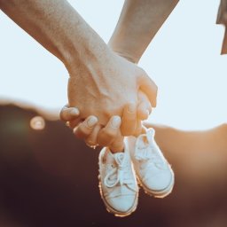 FQ Fotografia - Złączone dłonie pary trzymające białe, sznurowane buciki niemowlęce na tle rozmytego krajobrazu w ciepłych, zachodzących promieniach słońca.