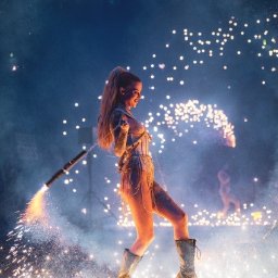 FQ Fotografia - Kobieta w kostiumie scenicznym, z wysokim kucykiem, trzyma w ręku urządzenie wyrzucające iskry, na scenie w otoczeniu efektów pirotechnicznych.